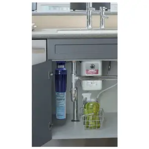 An open cabinet door under a kitchen sink showcases a water filtration system installed with thoughtful plumbing design. The setup includes a blue cylindrical filter connected to a white unit. A basket containing two green bottles is conveniently placed at the bottom right corner of the cabinet.