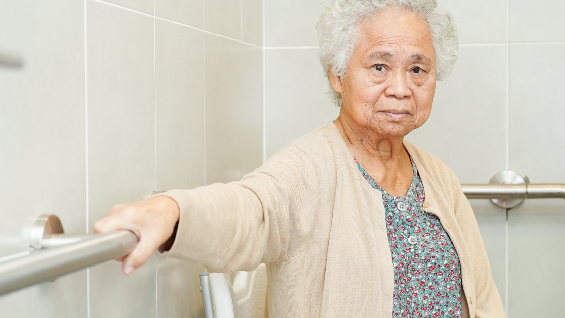An elderly Asian woman with gray hair, wearing a cardigan and floral top, holding onto a grab bar in a bathroom, looking at the camera with a composed expression near some plumbing fixtures.