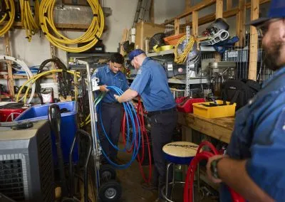 Two technicians in blue uniforms working with equipment in a cluttered workshop